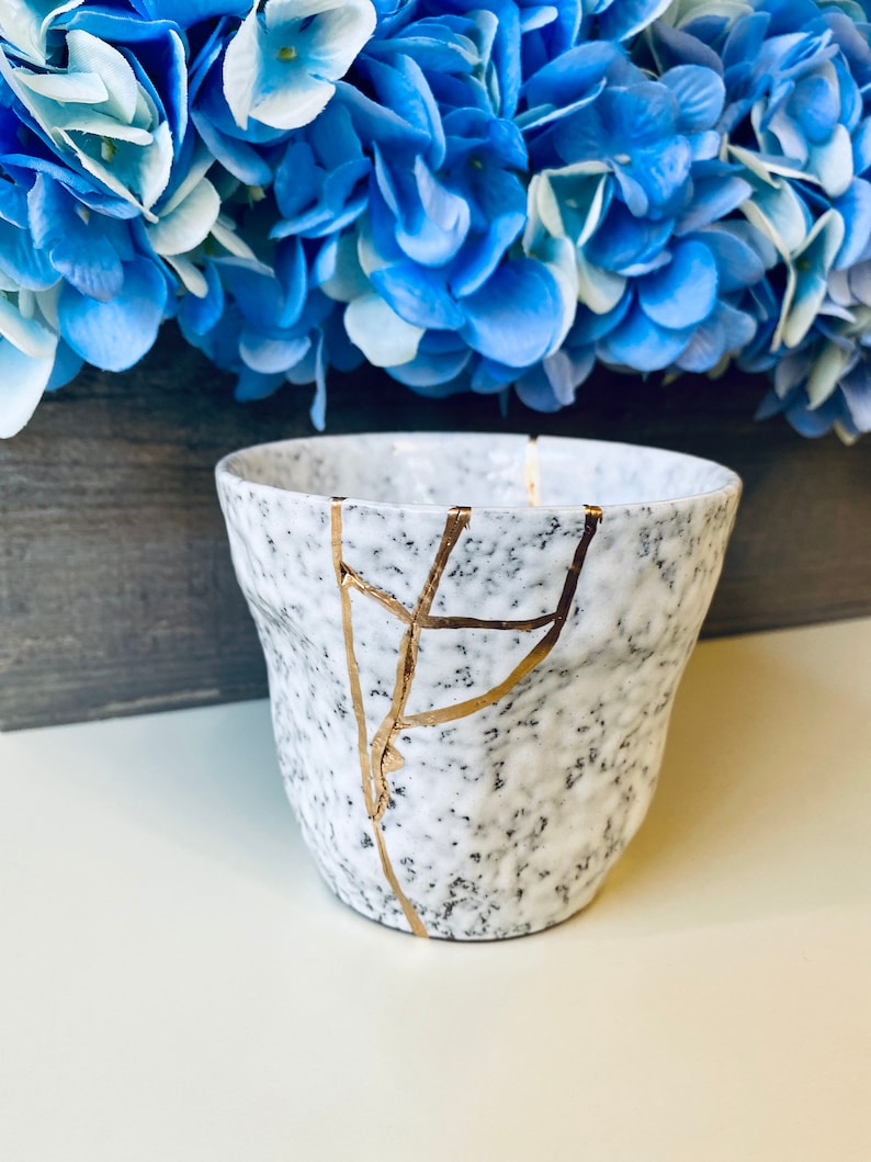 Pode incluir: Caneca de cer&acirc;mica branca com acabamento salpicado e design de rachaduras douradas. A caneca est&aacute; sobre uma superf&iacute;cie branca.