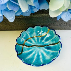 Può includere: Un piccolo piatto in ceramica blu e bianco con un bordo a forma di conchiglia e dettagli dorati. Il piatto ha una smaltatura craquelé e una linea dorata che attraversa il centro.
