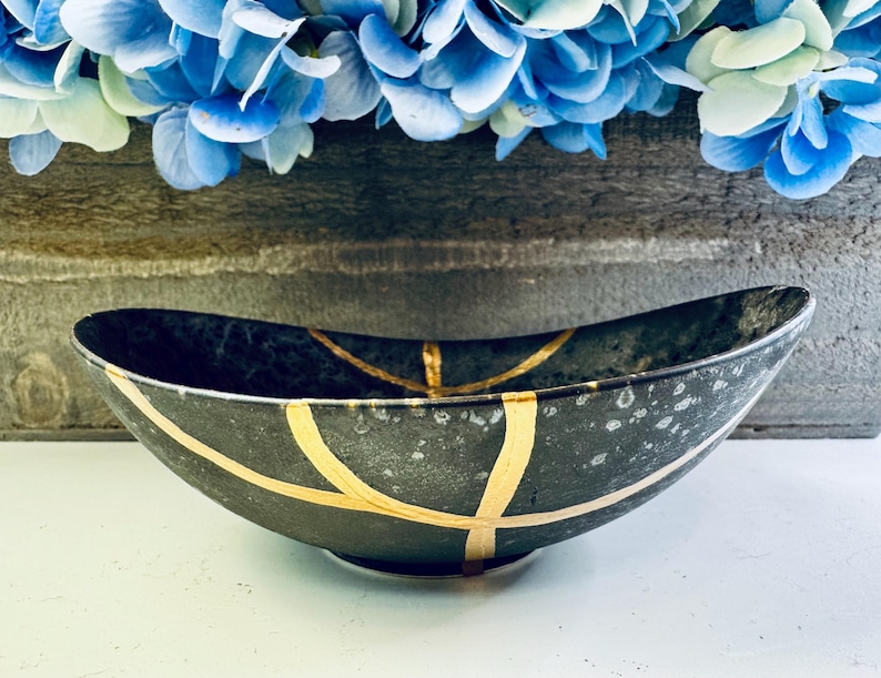 May include: A black oval bowl with gold accents. The bowl has a speckled finish and is decorated with gold lines.