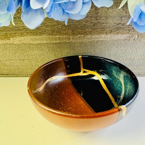 May include: A ceramic bowl with a unique Kintsugi repair. The bowl is divided into sections of brown and dark teal, with gold-colored lines highlighting the repaired cracks. The bowl is set against a neutral background.