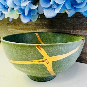 May include: A green ceramic bowl with gold-colored kintsugi repair lines. The bowl has a textured surface and a round base. Blue flowers are visible in the background.