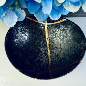 May include: A black ceramic bowl with gold accents. The bowl has a textured surface and a slightly rounded shape.