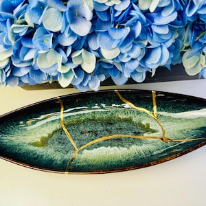Può includere: Un piatto decorativo ovale con un design marmorizzato verde scuro e bianco, accentuato da linee dorate. Il piatto è posto sotto un gruppo di fiori blu e bianchi.