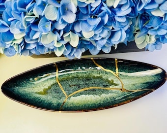 May include: An oval-shaped decorative dish with a dark green and white marbled design, accented with gold-colored lines. The dish is placed beneath a cluster of blue and white flowers.