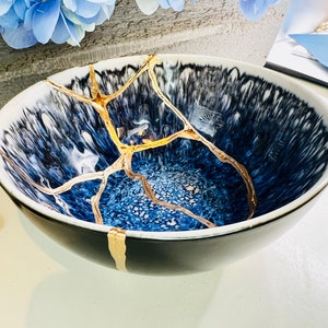 May include: A black and white ceramic bowl with a blue interior, featuring gold-colored kintsugi repairs. The bowl has a wide rim and a rounded shape. The kintsugi repairs are visible as golden lines across the surface.