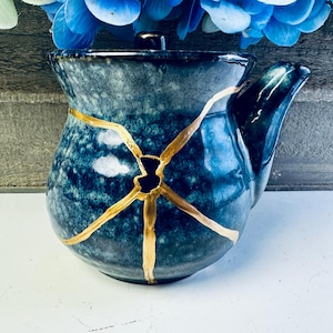 May include: A ceramic teapot with a dark blue and black mottled glaze, featuring a prominent gold-coloured repair line across the centre. The teapot has a rounded body, a short spout on the right side, and a matching lid with a small handle on top.