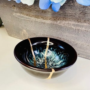 May include: A small, black ceramic bowl with a blue and white speckled glaze. The bowl has gold accents around the rim and on the inside.