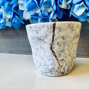 Pode incluir: Uma caneca de cer&acirc;mica branca com uma superf&iacute;cie texturizada e um design de rachadura dourada. A caneca est&aacute; sobre uma superf&iacute;cie branca.
