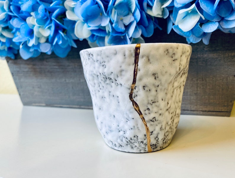 Pode incluir: Uma caneca de cer&acirc;mica branca com uma superf&iacute;cie texturizada e um design de rachadura dourada. A caneca est&aacute; sobre uma superf&iacute;cie branca.