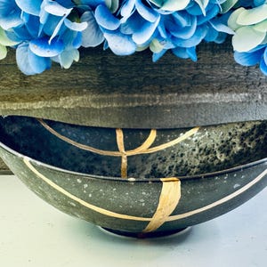May include: A black oval bowl with gold lines and a speckled finish. The bowl is sitting on a white surface.