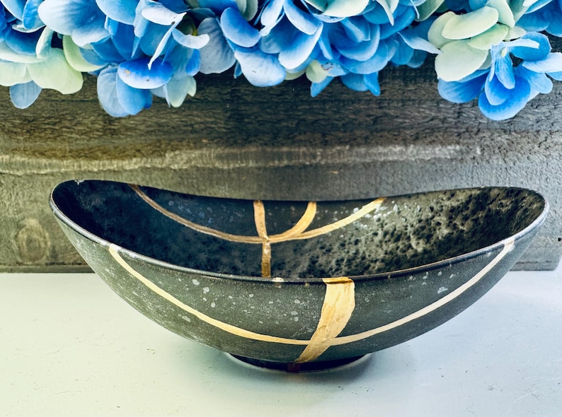 May include: A black oval bowl with gold lines and a speckled finish. The bowl is sitting on a white surface.