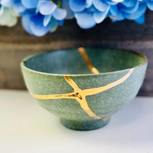Puede incluir: Un cuenco de cerámica verde celadón con líneas doradas de reparación kintsugi. El cuenco tiene forma redondeada y una pequeña base con pie. El fondo presenta flores azules y una superficie de madera.