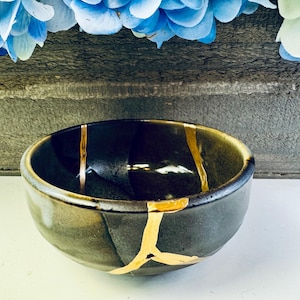 Puede incluir: Un cuenco de cerámica gris oscuro con líneas de reparación kintsugi doradas. El cuenco tiene forma redondeada y un acabado brillante. Flores azules visibles en el fondo.
