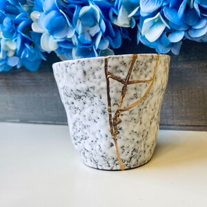 Pode incluir: Caneca de cer&acirc;mica branca com superf&iacute;cie texturizada e design de rachaduras douradas. A caneca est&aacute; ligeiramente lascada.