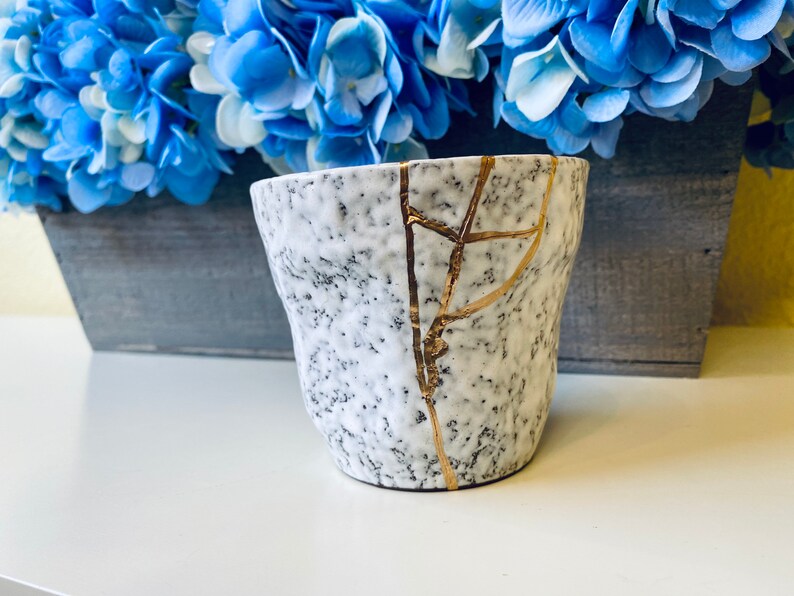 Pode incluir: Caneca de cer&acirc;mica branca com superf&iacute;cie texturizada e design de rachaduras douradas. A caneca est&aacute; ligeiramente lascada.
