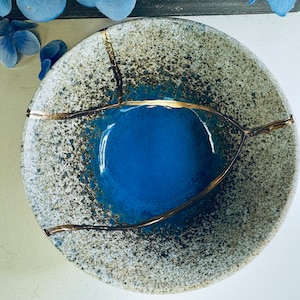 Op de afbeelding: Een blauwe keramische kom met een gespikkelde glazuur en gouden accenten. De kom heeft een gekrakelde afwerking en een uniek ontwerp.