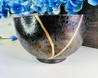 May include: A black ceramic bowl with gold accents. The bowl has a textured surface and a wide, round shape.