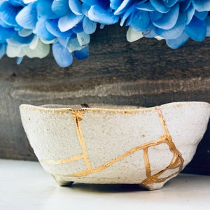 Puede incluir: Un cuenco de cerámica blanca con grietas doradas. El cuenco es ligeramente ovalado y tiene una superficie texturizada.