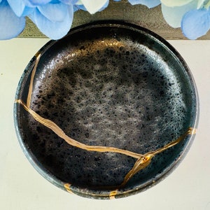 Puede incluir: Un cuenco redondo de cerámica negra con una textura moteada y una reparación kintsugi dorada. El cuenco tiene un acabado oscuro y brillante y una profundidad baja. Los detalles dorados resaltan las grietas, añadiendo un elemento decorativo.