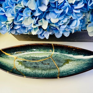 Puede incluir: Un plato decorativo ovalado de cerámica con borde marrón oscuro e interior verde azulado y blanco. El plato presenta reparaciones kintsugi doradas. Flores de hortensia azules en el fondo.
