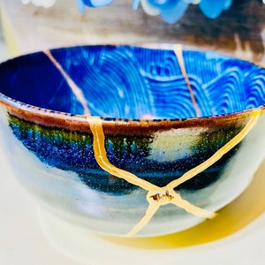 Puede incluir: Un cuenco de cerámica azul y marrón con grietas doradas. El cuenco tiene un interior azul texturizado y un borde marrón.