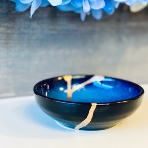 May include: A small, blue and black ceramic dish with gold accents. The dish has a crackled glaze and a rounded shape.
