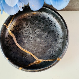 May include: A black ceramic bowl with gold crackle lines. The bowl has a textured surface and is ideal for serving small snacks or holding jewellery.