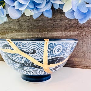 May include: A blue and white ceramic bowl with a wave pattern and gold accents, showcasing the Japanese art of Kintsugi. The bowl is set against a wooden background with blue flowers.