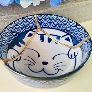Puede incluir: Un pequeño cuenco de cerámica azul y blanco con un borde dorado y un diseño de gato. El cuenco está agrietado y reparado con kintsugi dorado.