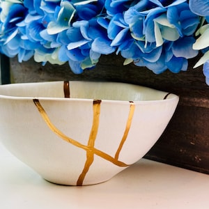 May include: A white ceramic bowl with a unique Kintsugi design, featuring gold-colored lines that mend the cracks. The bowl has a slightly asymmetrical shape and is set against a backdrop of blue flowers.