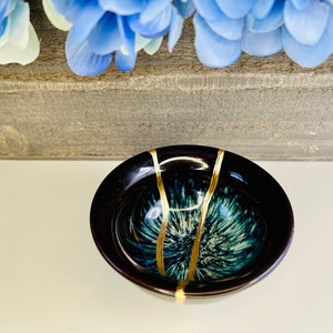 Może przedstawiać: Mała, czarna ceramiczna miseczka z wzorem w postaci niebiesko-zielonego i białego wiru oraz złotymi akcentami. Miseczka idealnie nadaje się do serwowania małych przekąsek lub dipów.