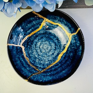 Può includere: Un piatto rotondo in ceramica blu scuro con un motivo a vortice strutturato. Il piatto presenta linee color oro che evidenziano le crepe, mostrando l'arte giapponese del Kintsugi. Il bordo è di colore blu scuro.