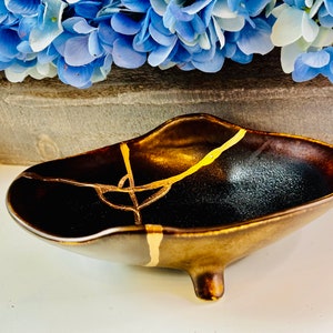 May include: A small, dark brown ceramic dish with a crackled glaze and gold accents. The dish has three small feet and is slightly curved.