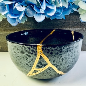 May include: A black and gray ceramic bowl with gold-colored kintsugi repair lines. The bowl has a speckled pattern and a smooth, glossy finish. Blue flowers are in the background.