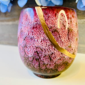 May include: A small, round, red ceramic cup with a white and gold design. The cup has a textured surface and a gold stripe around the rim.