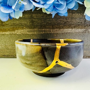 May include: A ceramic bowl with a dark, mottled exterior and gold-colored kintsugi repairs. The bowl has a rounded shape and sits on a small, dark base. Blue and white flowers are in the background.