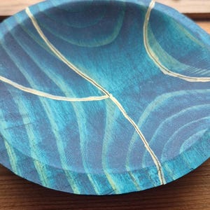 May include: A round, wooden decorative plate with a blue and teal painted design. The plate features a pattern of curved lines and is accented with thin, gold-colored lines. The plate is sitting on a wooden surface.