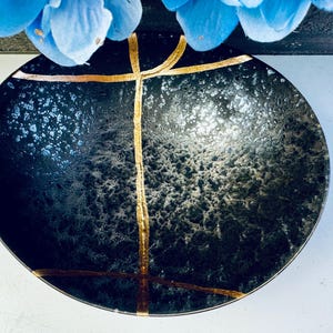 May include: A black ceramic bowl with a speckled finish and gold accents. The bowl has a wide rim and a rounded bottom.