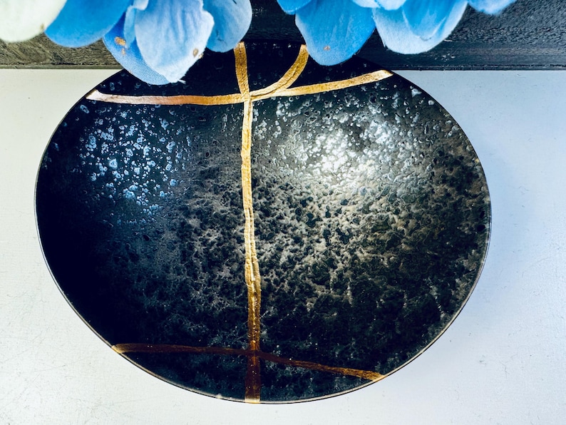 May include: A black ceramic bowl with a speckled finish and gold accents. The bowl has a wide rim and a rounded bottom.