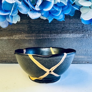 Può includere: Una ciotola in ceramica nera con bordo smerlato, caratterizzata da linee di riparazione kintsugi color oro. La ciotola è ambientata su uno sfondo di fiori blu e una superficie grigio scuro, mostrando un mix di design tradizionale e moderno.