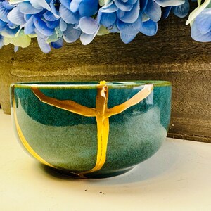 May include: A teal ceramic bowl with gold accents. The bowl has a rounded shape and a smooth, glossy finish.