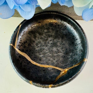May include: A black ceramic dish with gold crackle lines. The dish is round and has a slightly raised rim.