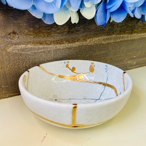 May include: A small white ceramic bowl with a gold rim and delicate blue and gold floral designs. The bowl is perfect for serving small snacks or dips.