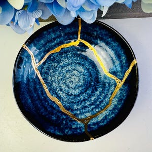 May include: A round, dark blue ceramic bowl with a speckled pattern and gold-coloured kintsugi repair lines. The bowl has a black rim and is set against a white background, with blue flowers in the upper portion of the image.