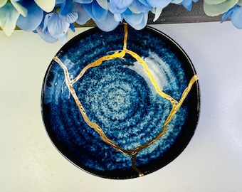Puede incluir: Un cuenco redondo de cerámica azul oscuro con un patrón moteado y líneas de reparación kintsugi doradas. El cuenco tiene un borde negro y está sobre un fondo blanco, con flores azules en la parte superior de la imagen.