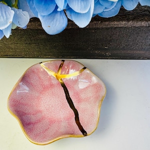 Può includere: Un piccolo piatto in ceramica rosa di forma irregolare con dettagli dorati e una linea marrone scuro. Il piatto ha un bordo dorato e una crepa riparata con oro. Fiori blu visibili sullo sfondo.