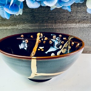 Op de afbeelding: Een keramische kom met een donkerblauwe binnenkant en een grijze buitenkant. De kom heeft goudkleurige kintsugi-reparaties en decoratieve accenten. De rand is warmbruin. Een uniek kunstwerk.