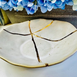 Könnte beinhalten: Eine weiße Keramikschüssel mit craquelé-Glasur und goldenen Akzenten. Die Schüssel hat eine einzigartige, organische Form und ist perfekt zum Servieren von Snacks oder Desserts.