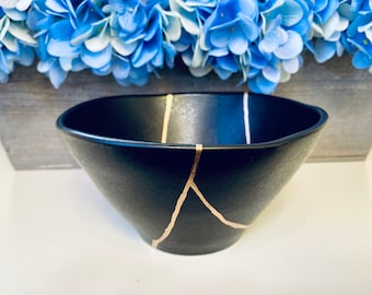 May include: A black ceramic bowl with gold crackle lines. The bowl is slightly oval shaped and has a wide rim.