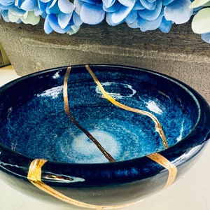 May include: A deep blue ceramic bowl with gold-colored kintsugi repair lines. The bowl has a glossy finish and a gradient effect, transitioning from dark blue to lighter shades. Decorative blue flowers are in the background.